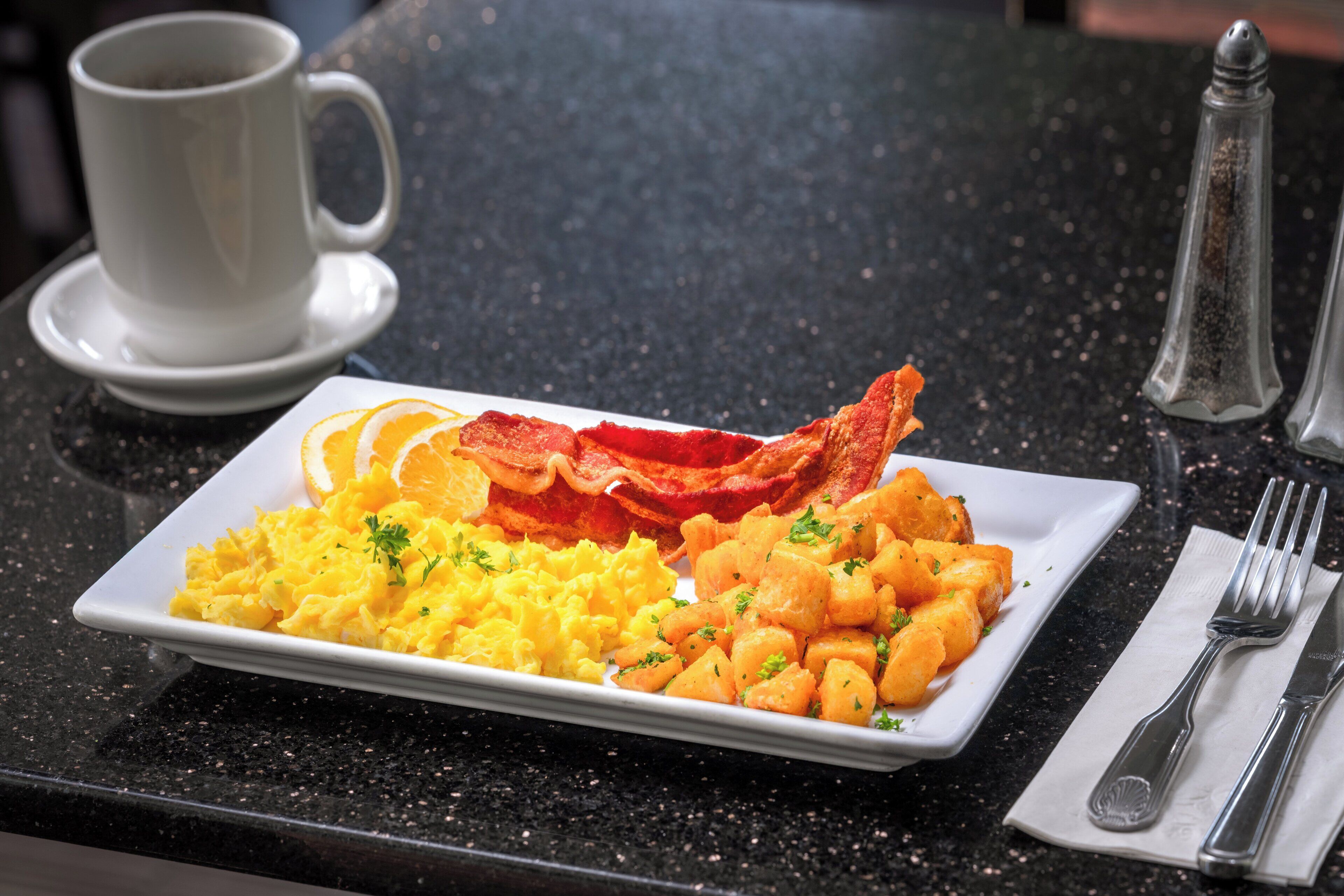 A breakfast plate with scrambled eggs, bacon, and hash browns on a black countertop.