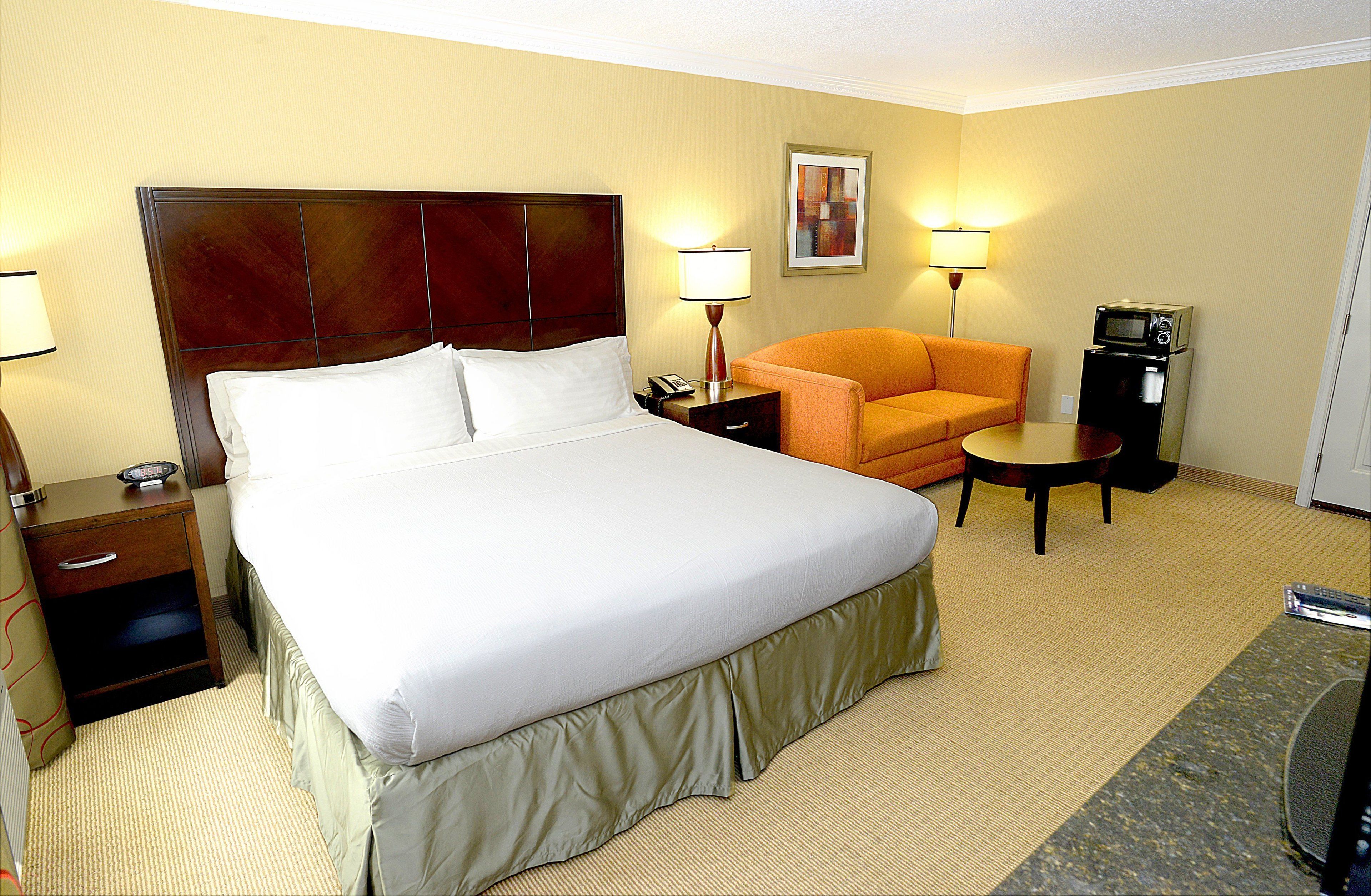 A hotel room with a large bed, a nightstand, a lamp, an orange chair, a coffee maker, and a small table.