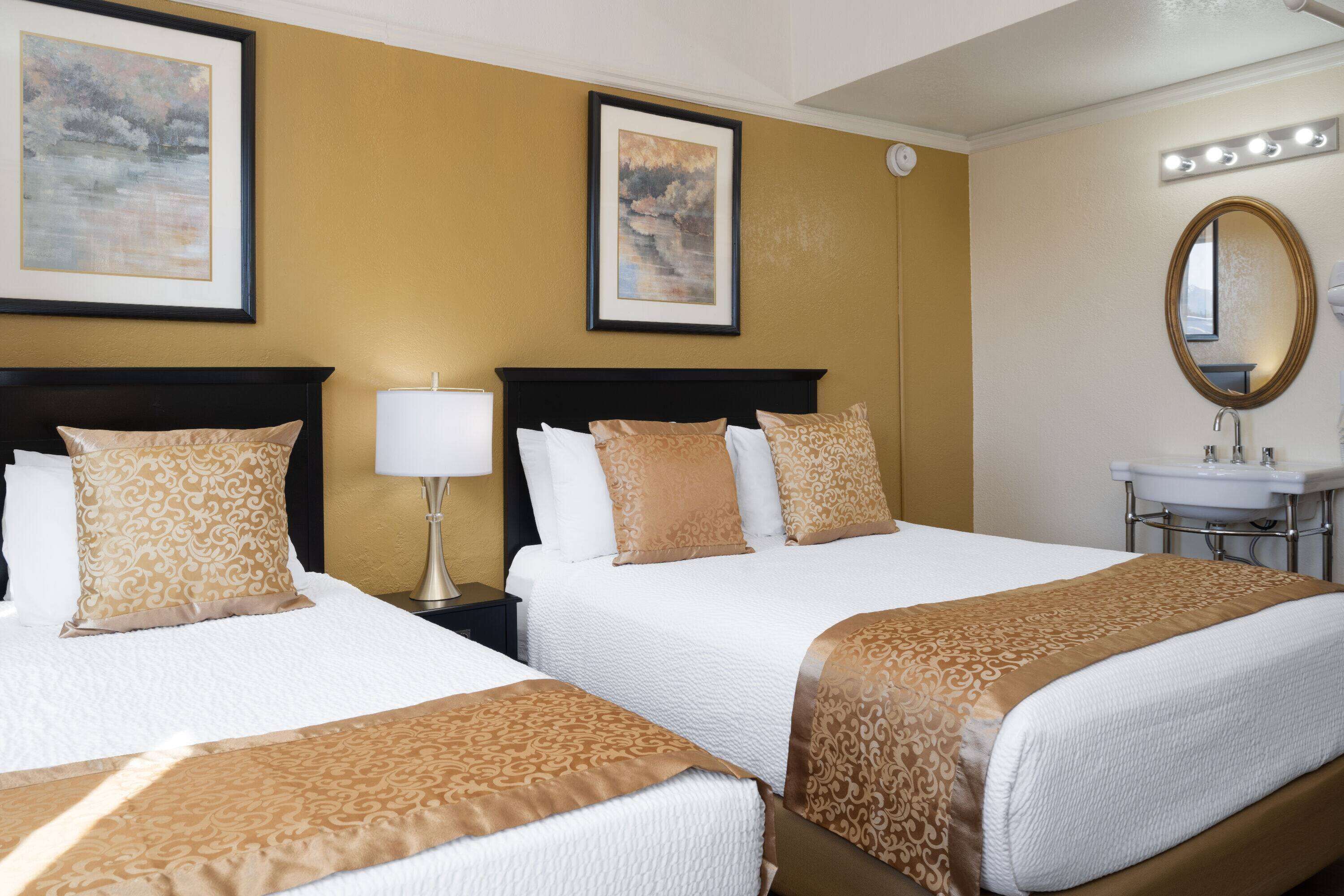 A hotel room with two beds, a bedside table with a lamp, a bathroom with a sink and mirror, and framed artwork on the walls.