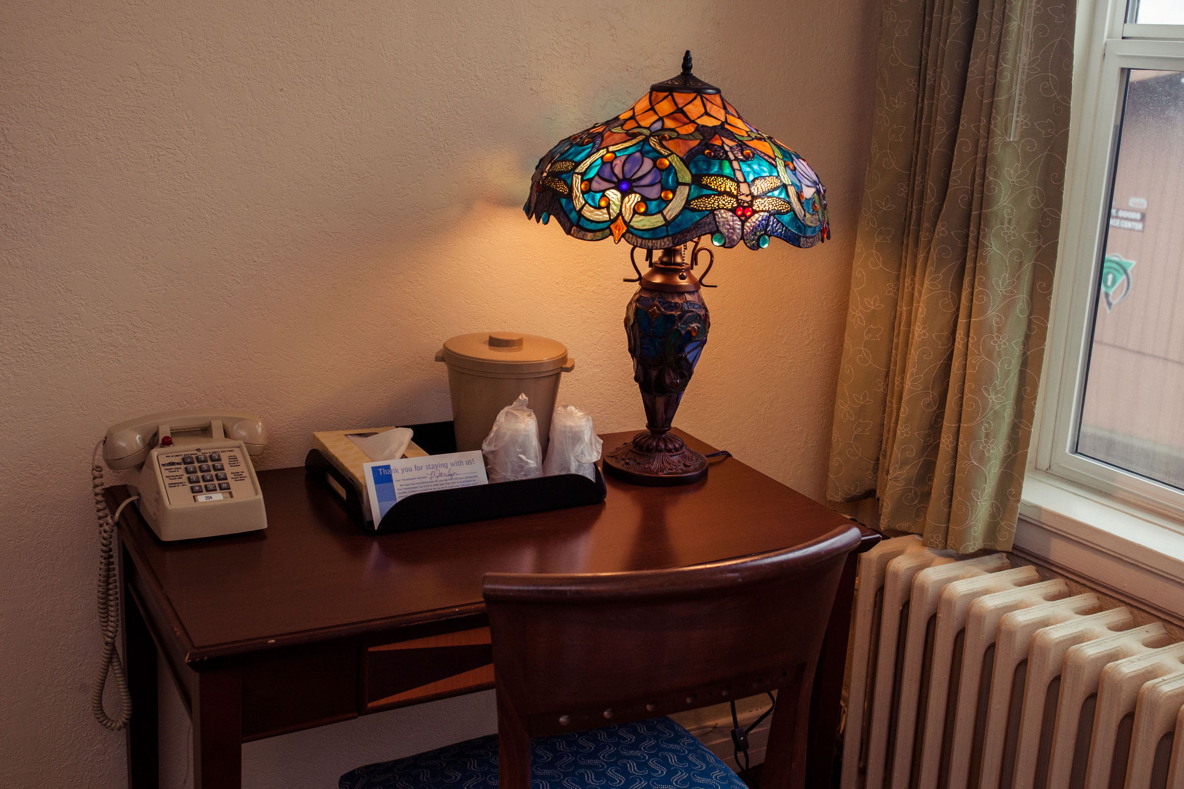 A hotel room with a wooden desk, a telephone, a decorative stained glass lamp, a tissue box, and a radiator.