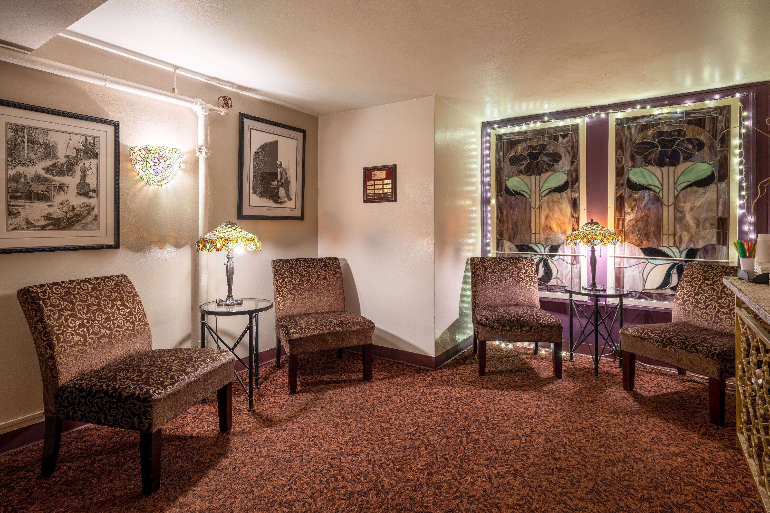 A room with patterned carpet, multiple chairs, a table with a lamp, framed pictures on the wall, and a decorative window with lights.