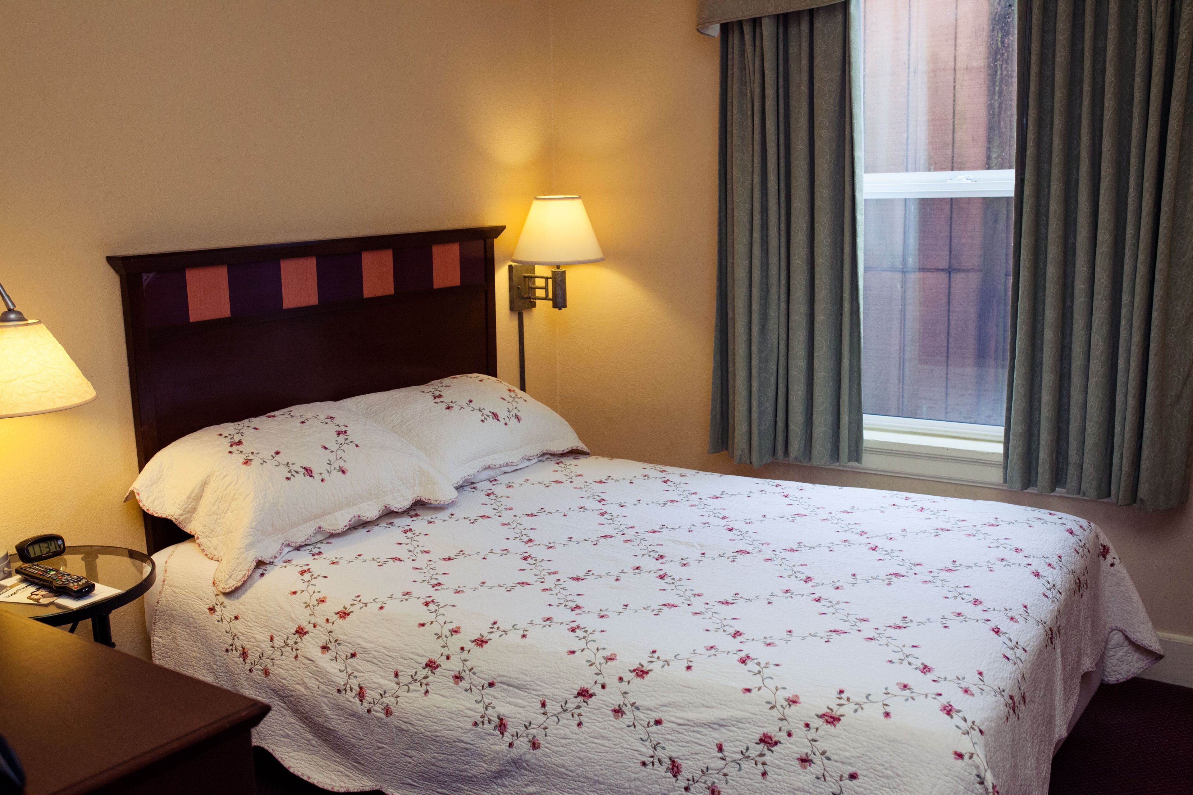 Una cama bien hecha con una colcha estampada, una mesita de noche con una lámpara y una ventana con cortinas.
