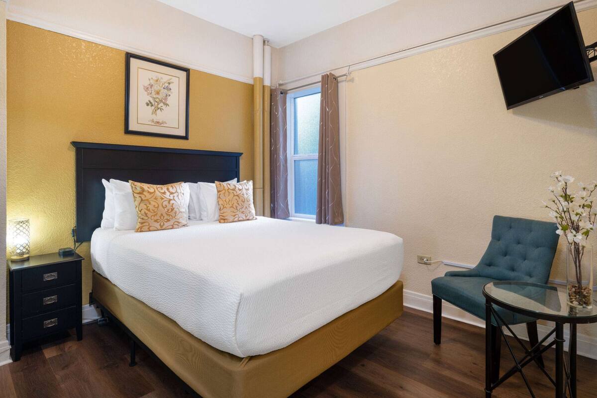 Un dormitorio ordenado con una cama grande, una mesita de noche, una silla y un televisor montado en la pared.
