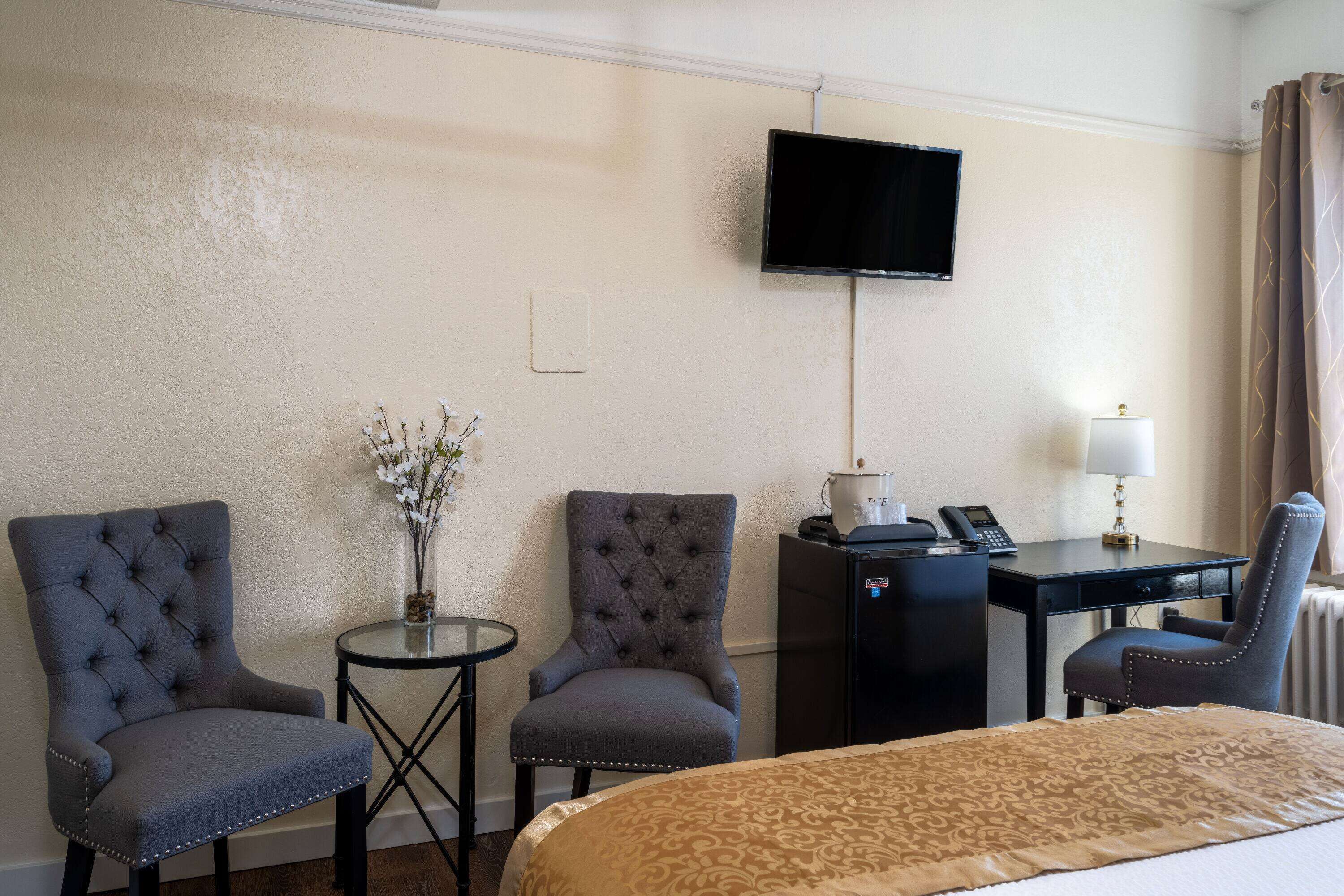 Ein Hotelzimmer mit einem Bett, zwei Sesseln, einem kleinen Tisch mit Lampe, einem Schreibtisch mit Telefon und einem Fernseher an der Wand.
