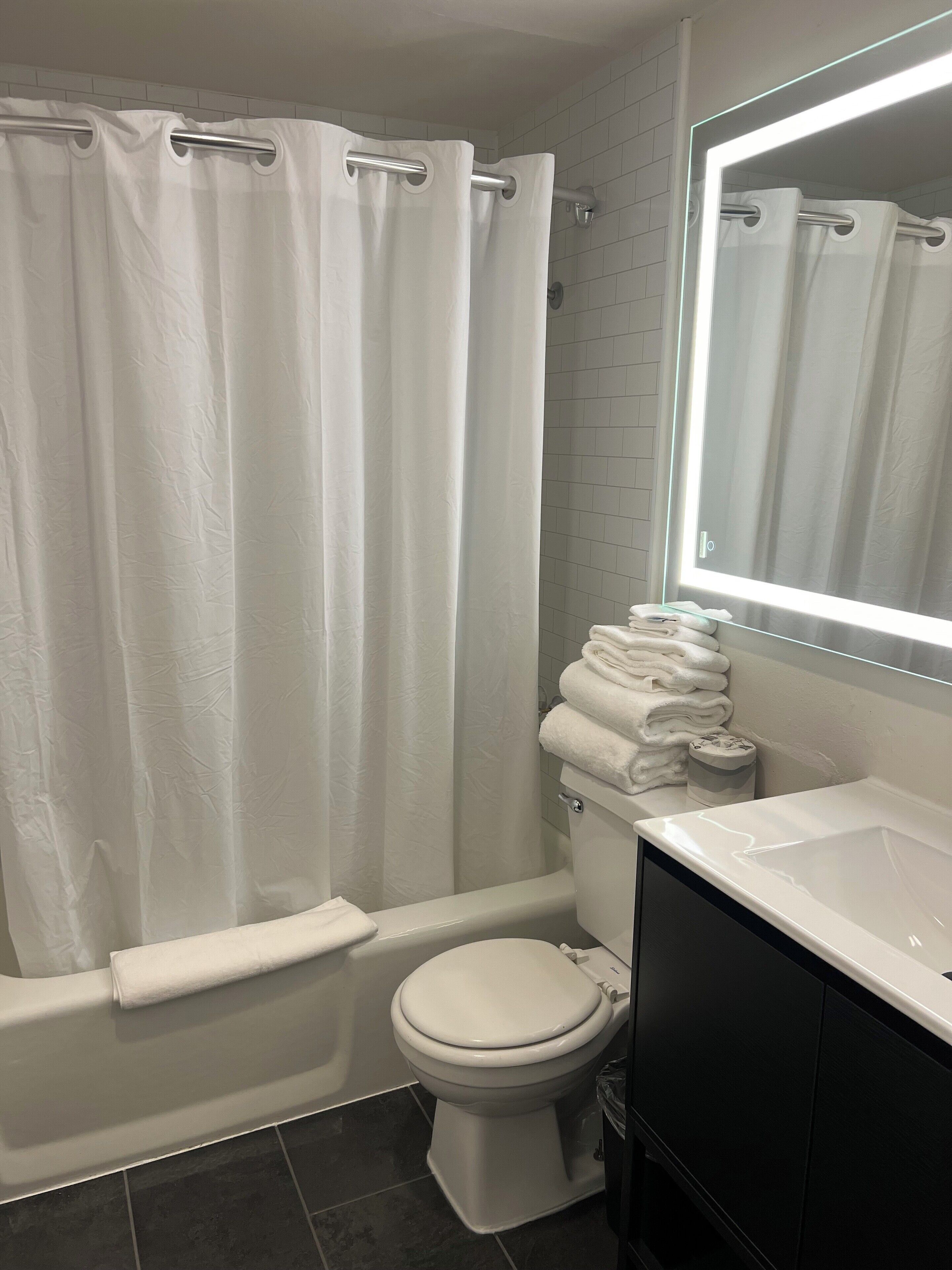 A bathroom with a white toilet, a bathtub with a white shower curtain, a sink, and a mirror with a light above it.
