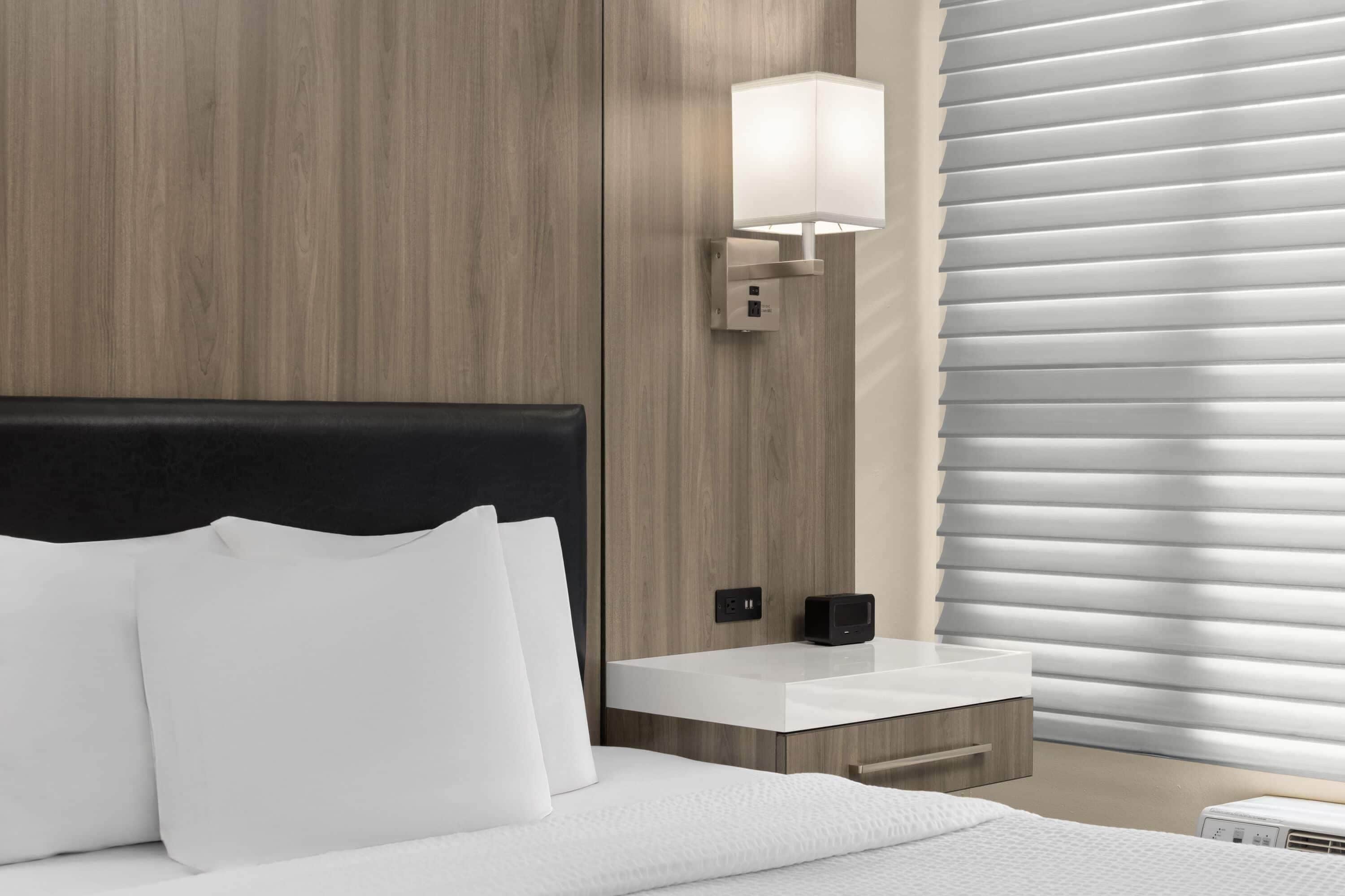 Una habitación de hotel moderna con cabecera de madera, una mesita de noche con lámpara y una ventana con persianas.