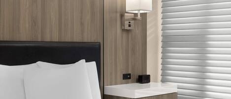 Una habitación de hotel moderna con cabecera de madera, una mesita de noche con lámpara y una ventana con persianas.