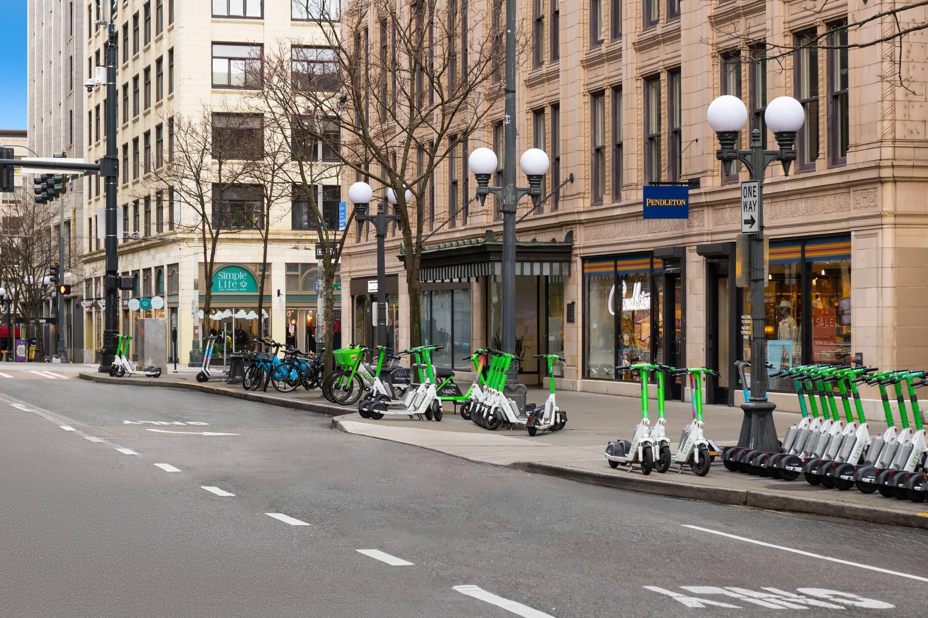 Une rue bordée d’une rangée de trottinettes électriques garées sur le trottoir, avec un magasin appelé « Simple Life » et un magasin « Pendleton ».
