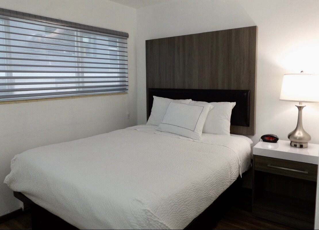 Un lit bien fait, avec du linge de lit blanc, une tête de lit en bois, une table de chevet avec une lampe et une fenêtre avec des stores.