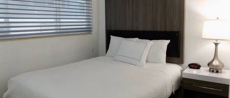 Un lit bien fait, avec du linge de lit blanc, une tête de lit en bois, une table de chevet avec une lampe et une fenêtre avec des stores.