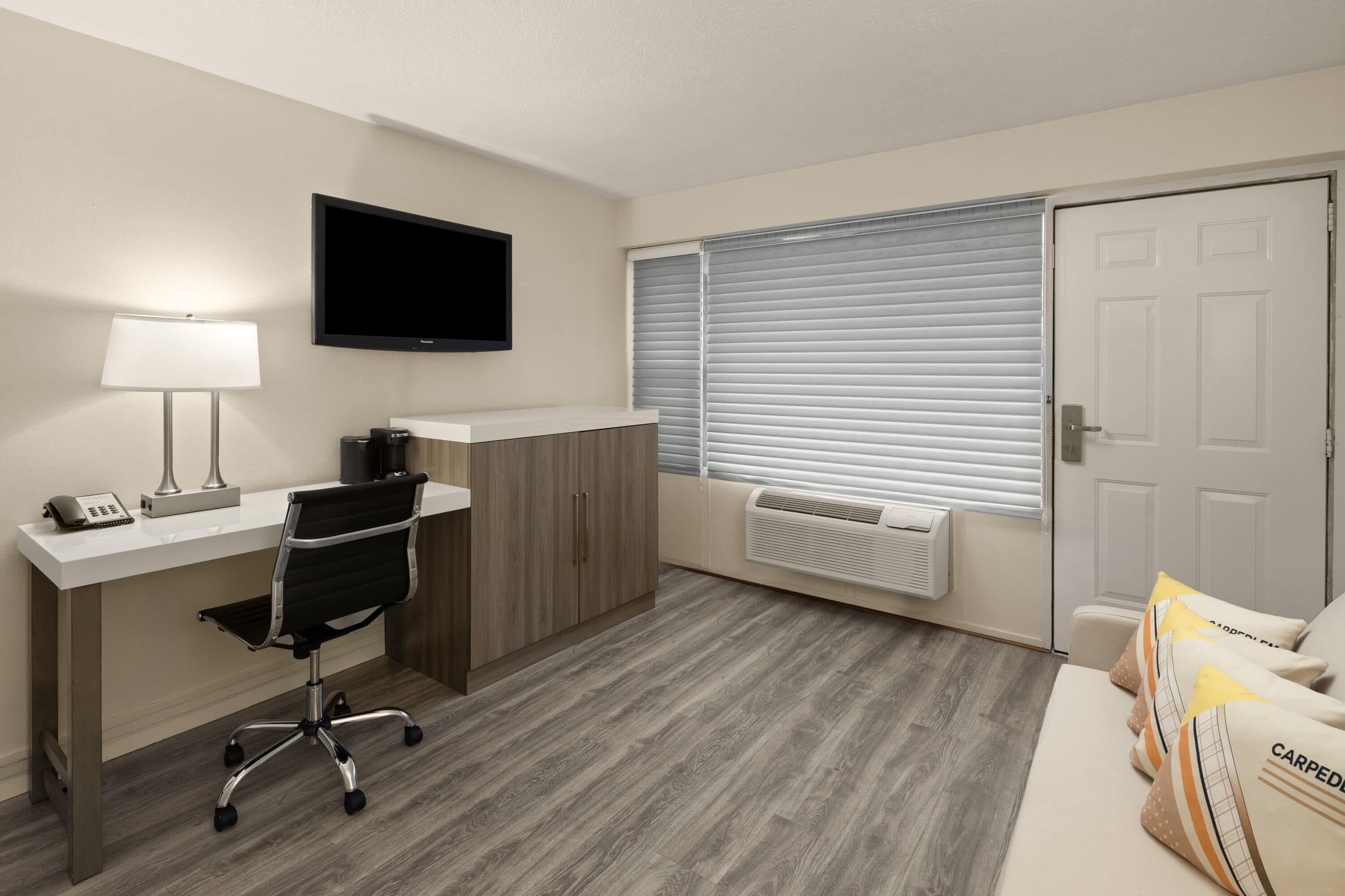 A room with a desk, chair, TV, window with blinds, and a door.