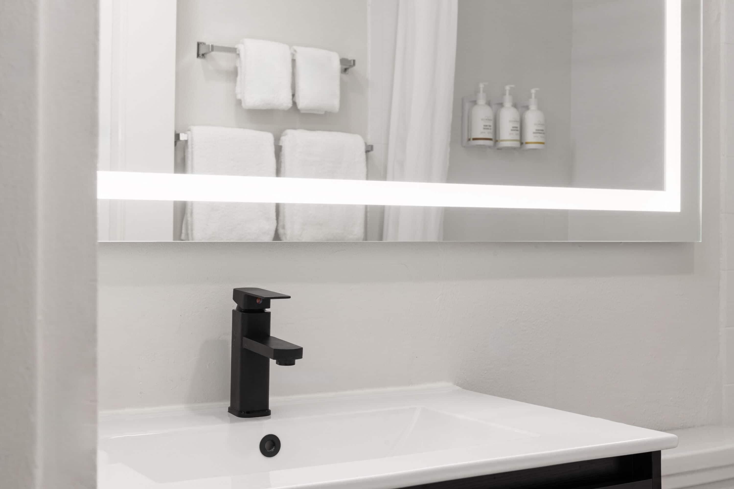 A bathroom with a white sink, black faucet, and a mirror reflecting a towel rack and toiletries.
