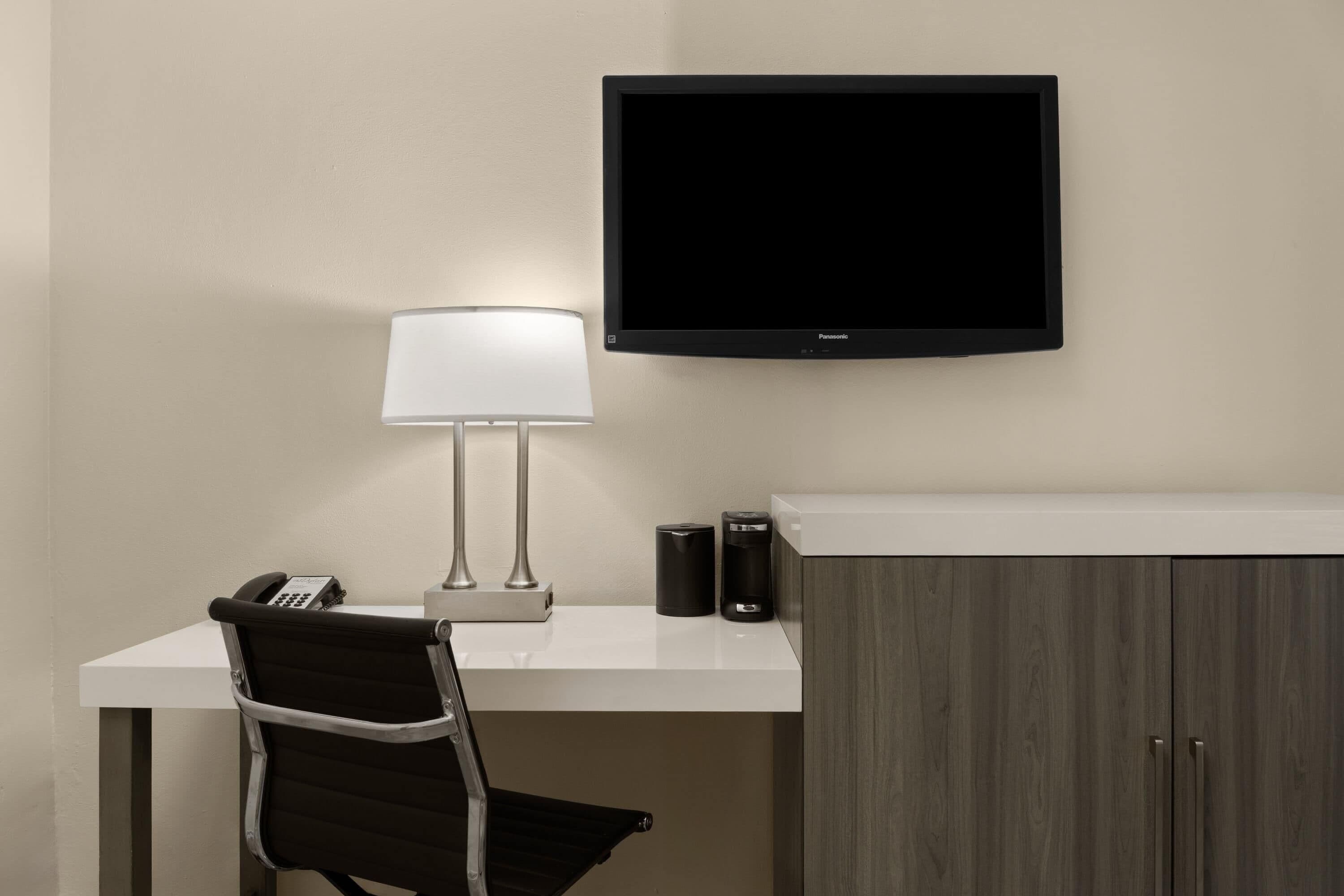 Et hotelværelse med skrivebord, stol, lampe, fladskærms-tv, telefon og skab.