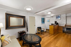 A living room with a glass-top coffee table, a sofa with cushions, a wooden chair, a desk with a computer, and a kitchen area with a counter and appliances.