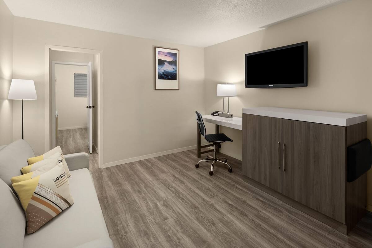 A hotel room with a sofa, a desk with a chair, a TV mounted on the wall, and a door leading to another room.