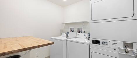 A laundry room with two washing machines, a drying rack, and a wooden table.