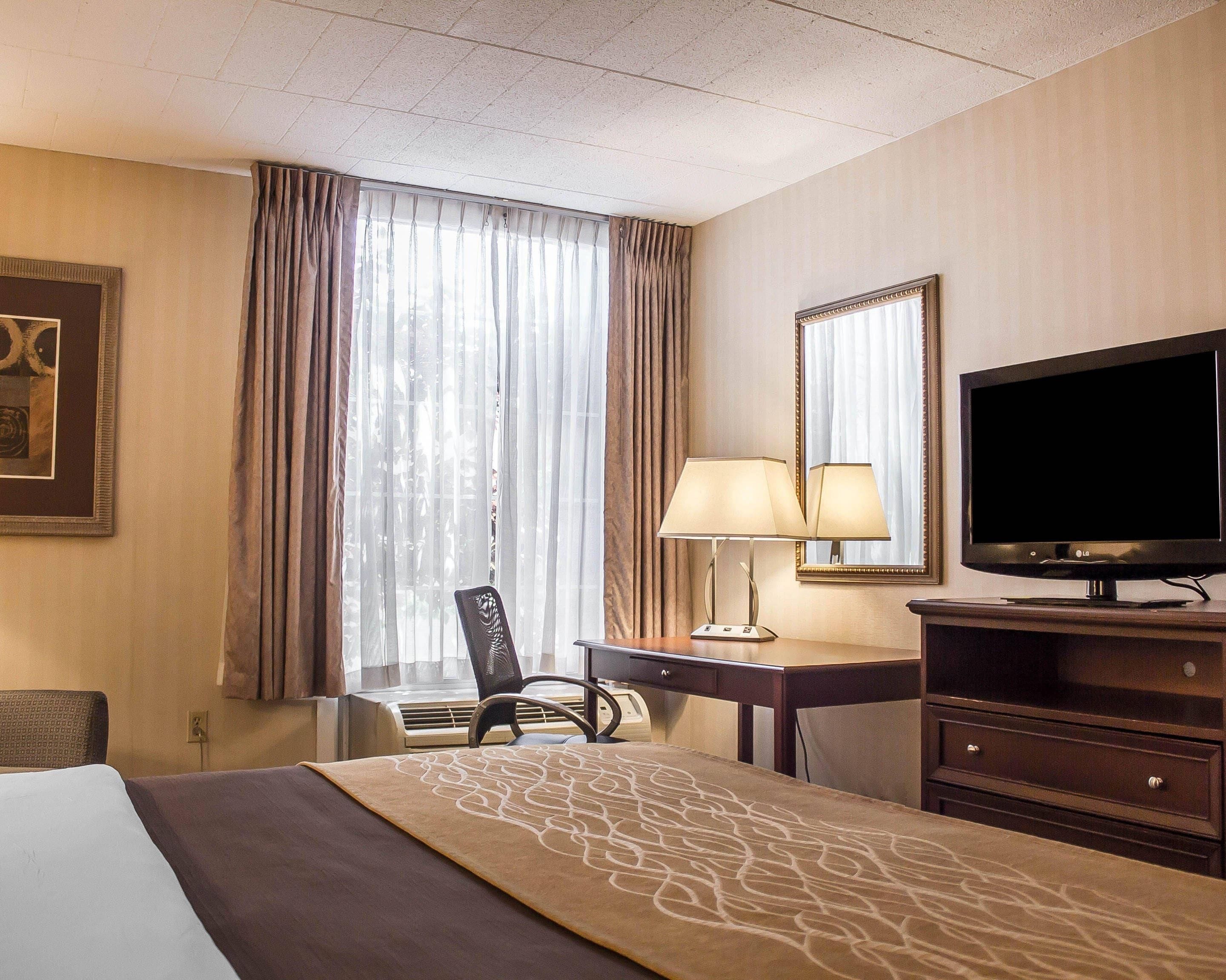 A hotel room with a bed, a desk with a lamp, a television, and a large window with curtains.