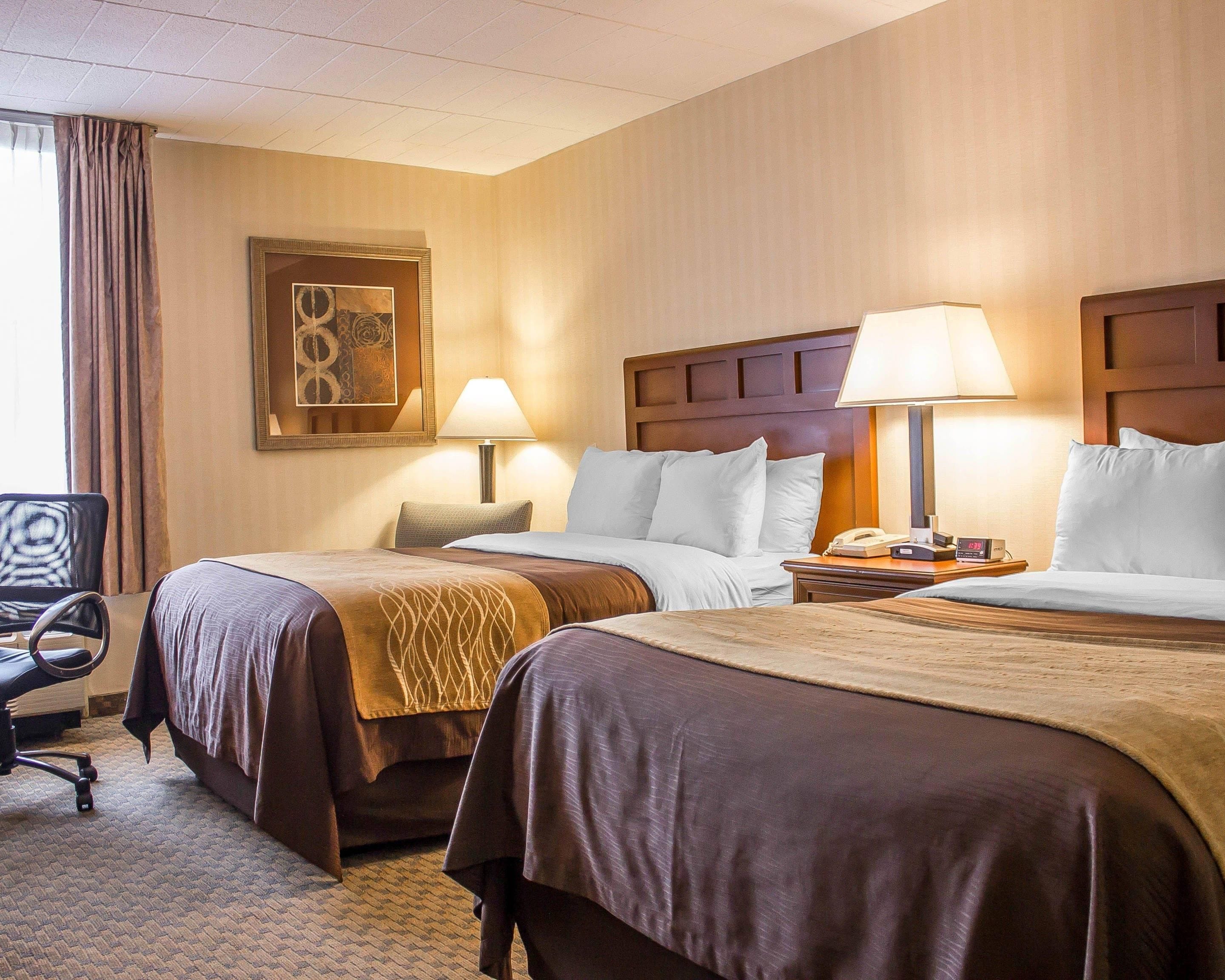 A hotel room with two beds, a desk, a chair, a lamp, a clock, and a framed picture on the wall.