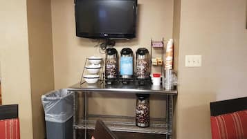 A hotel room with a flat-screen TV mounted on the wall, a coffee station with multiple coffee makers, and a trash bin.
