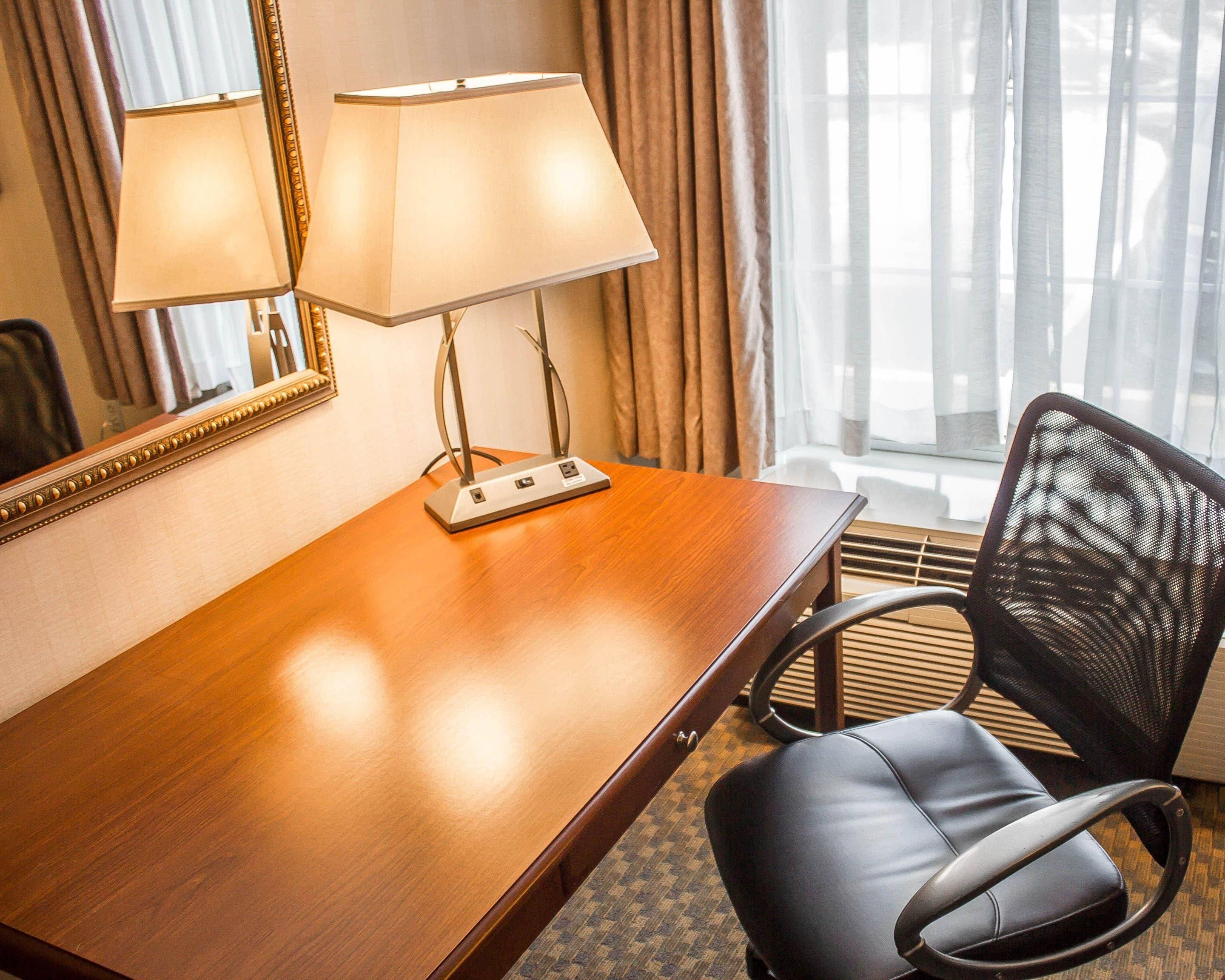 Habitación de hotel con escritorio de madera, dos lámparas, una silla, un espejo y una ventana con cortinas.