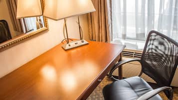Habitación de hotel con escritorio de madera, dos lámparas, una silla, un espejo y una ventana con cortinas.