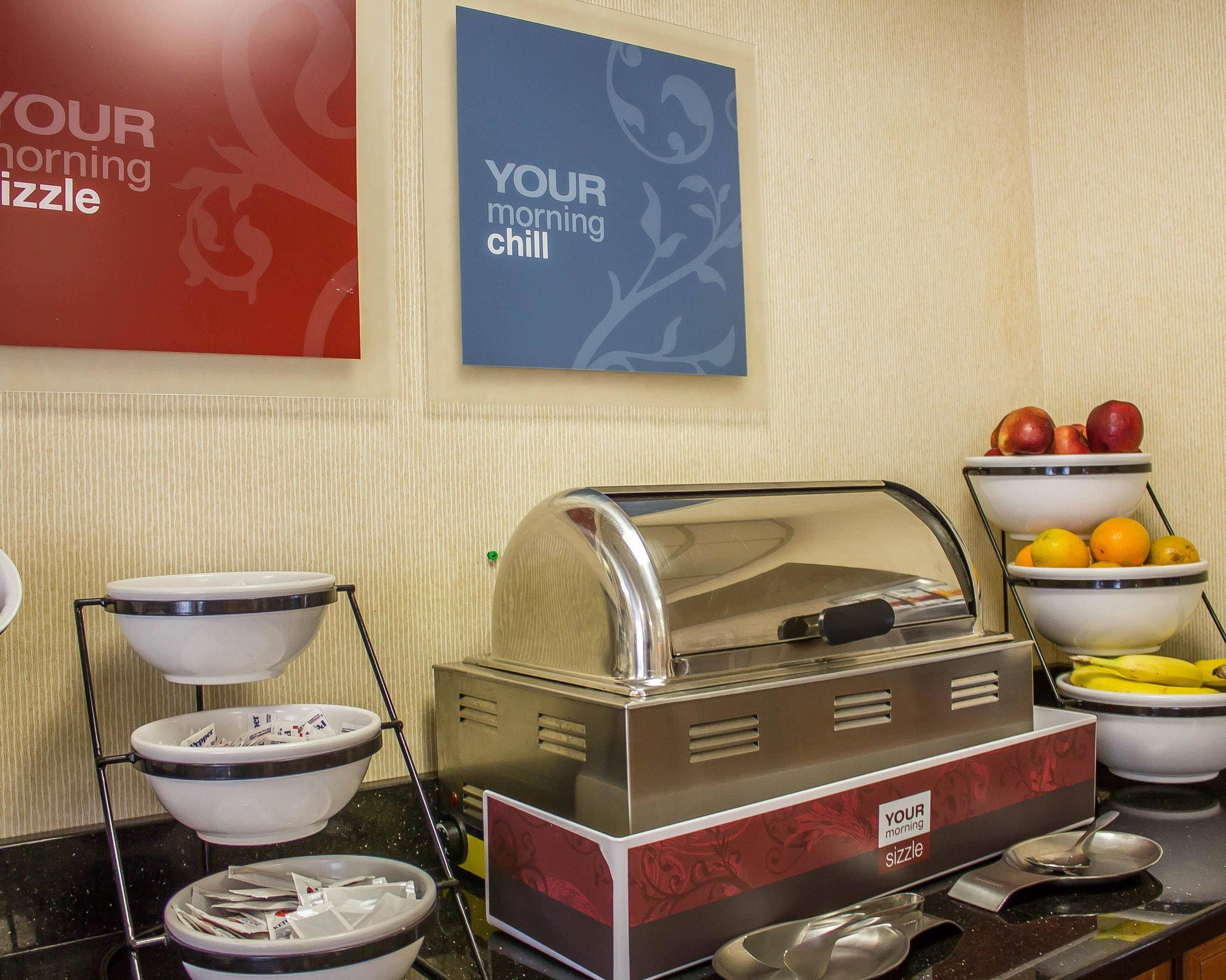 Montaje de desayuno buffet con una máquina de café, tazones de frutas y un letrero que dice 'YOUR morning sizzle'.