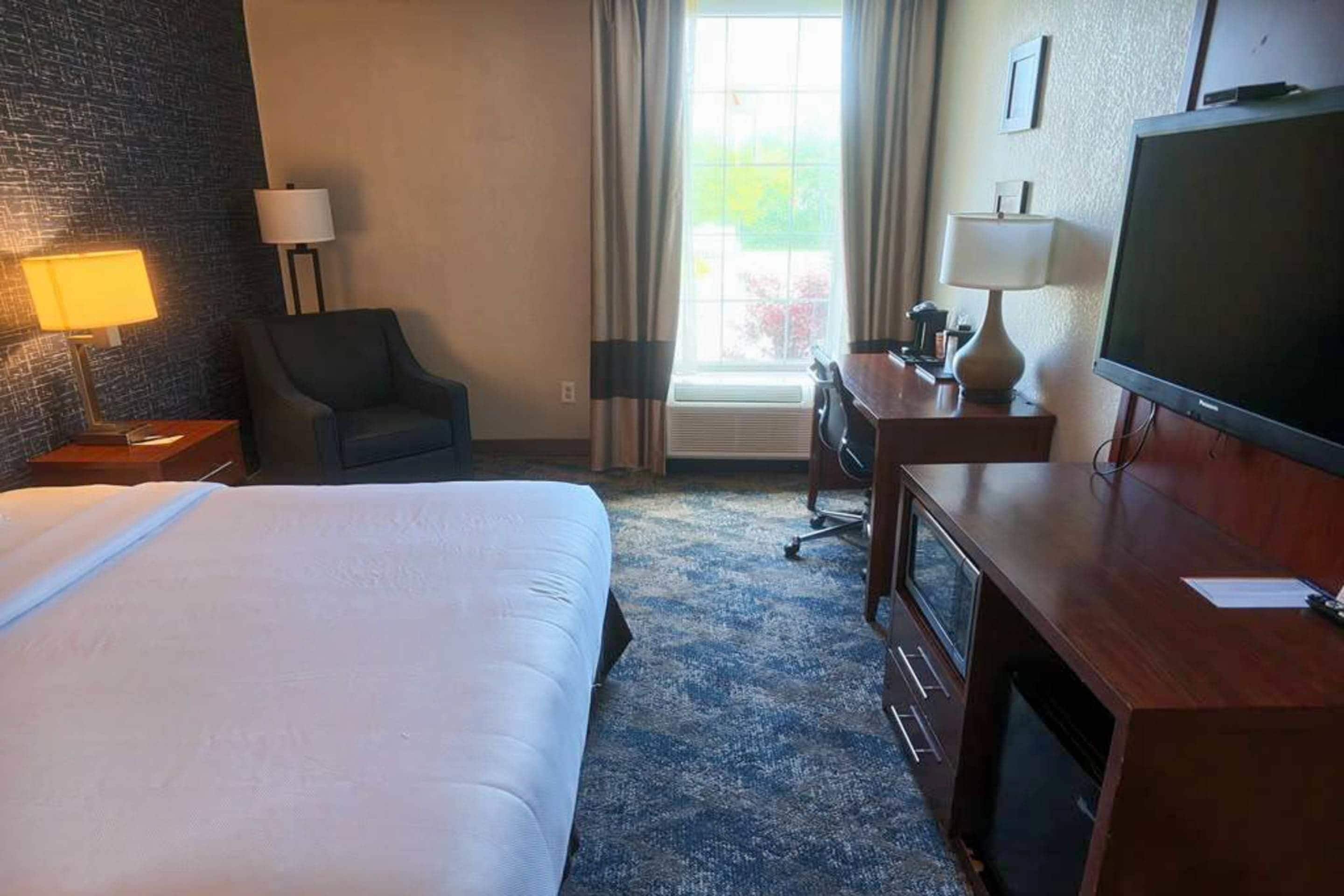 Habitación de hotel con cama, escritorio, silla, televisión y ventana con cortinas.