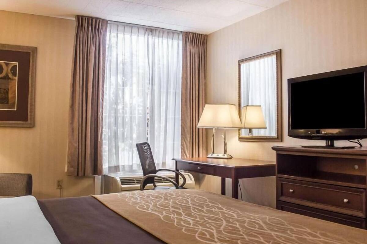 A hotel room with a bed, a desk with a lamp, a television, a chair, and a large window with curtains.