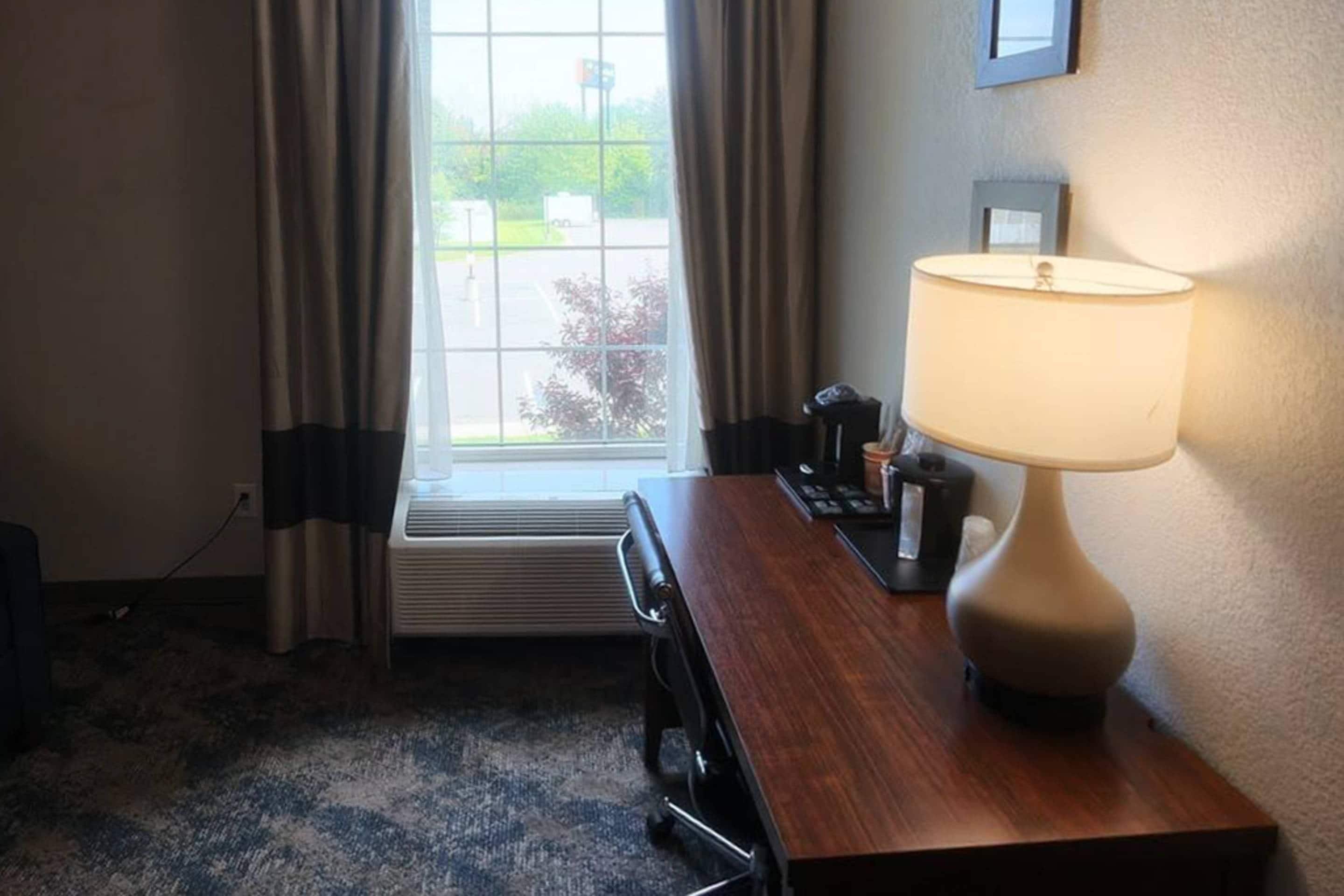 A hotel room with a desk, a lamp, a window with curtains, and a view of the outdoors.