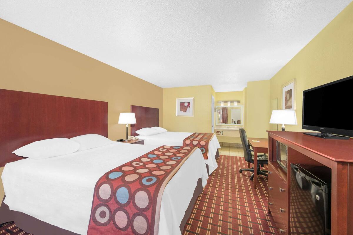 A hotel room with two beds, a desk, a television, and a bathroom visible in the background.