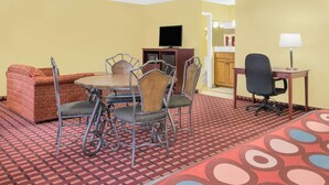 A hotel room with a dining table, four chairs, a sofa, a TV, a desk with a lamp, and a patterned carpet. - Super 8 by Wyndham Kerrville TX (Kerrville)