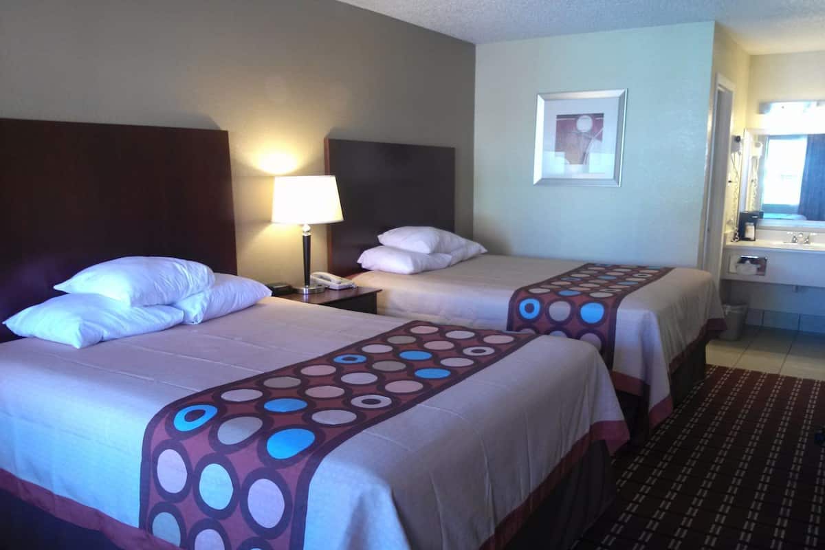 A hotel room with two beds, a nightstand with a lamp, a framed picture on the wall, and a bathroom with a sink and mirror.