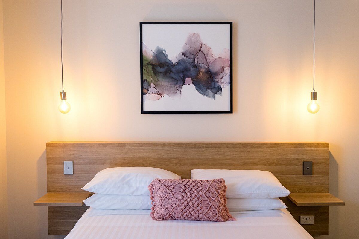 Una cama bien hecha con sábanas blancas y un cojín estampado en rosa, un cabecero de madera y un cuadro abstracto enmarcado encima.