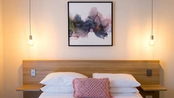 Un lit bien fait, avec du linge de lit blanc et un coussin à motifs roses, une tête de lit en bois et, au-dessus, un tableau abstrait encadré.