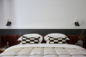 Una cama impecable con un cojín de cuadros, cabecero oscuro y dos lámparas de pared.