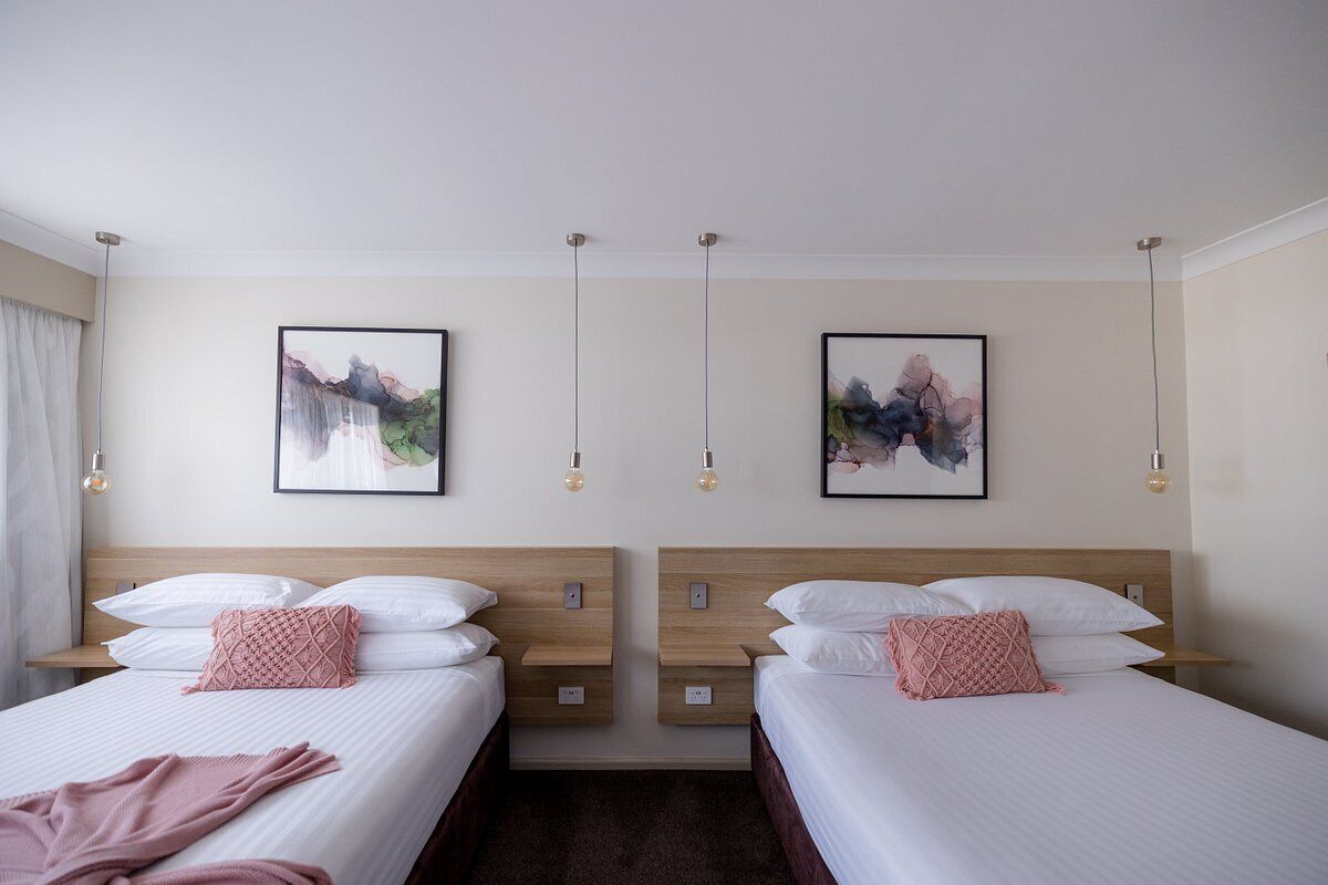 A hotel room with two beds, each with white linens and pink decorative pillows. There are two framed artworks on the wall, and pendant lights hanging above the beds.