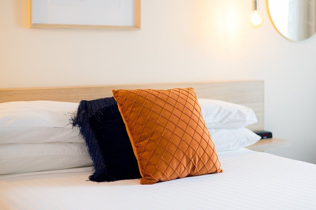 A neatly made bed with white and dark blue pillows, a wooden headboard, and a wall-mounted light fixture.