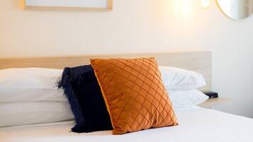 Una cama bien hecha con almohadas blancas y azul oscuro, un cabecero de madera y una lámpara fijada a la pared.