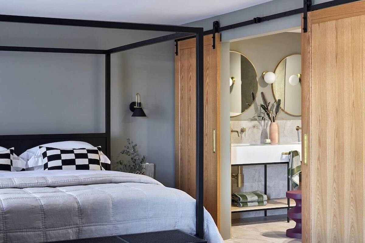 A modern bedroom with a bed, a bedside lamp, and a view into the bathroom through an open door.