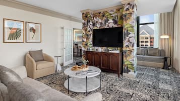 A modern living room with a sofa, armchair, and a round coffee table. A flat-screen TV is mounted on a wall with a floral mural. The room has a patterned carpet and a view of a building outside.