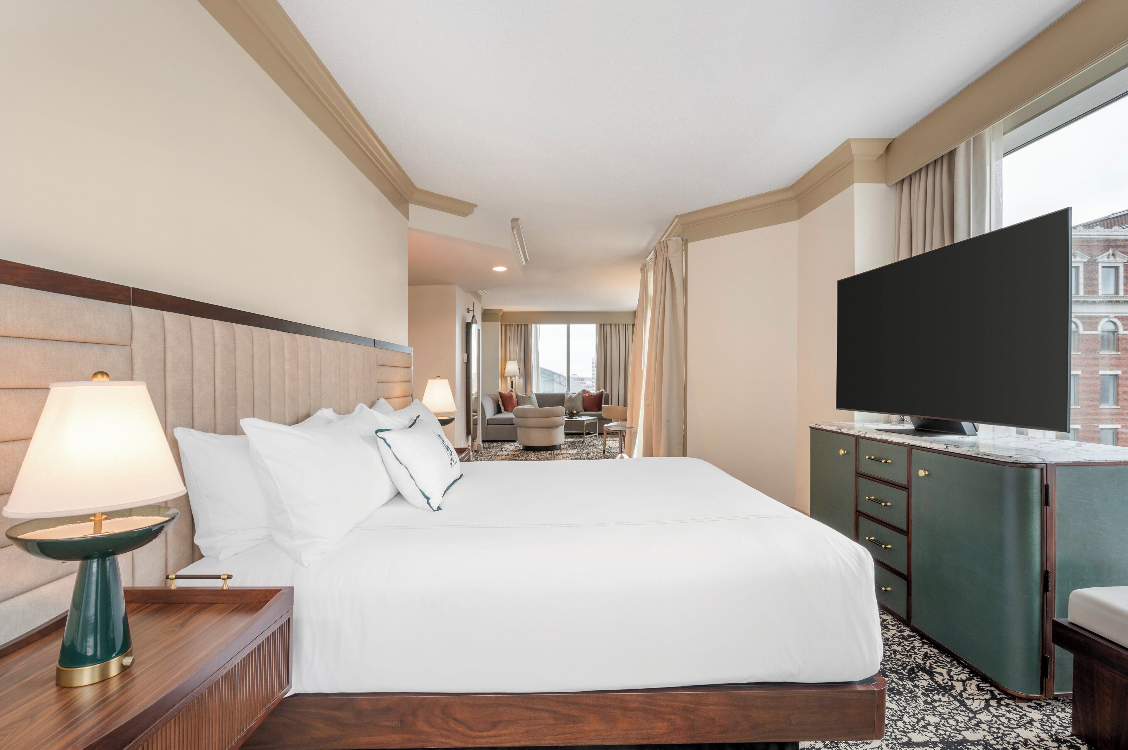 A hotel room with a large bed, a TV, a desk with a lamp, and a view of a building through the window.