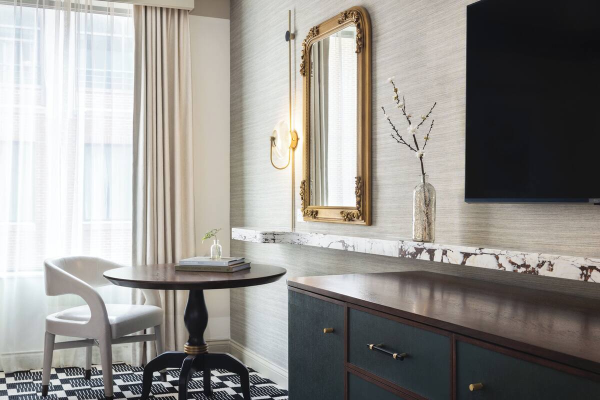 A modern hotel room with a marble wall, a large mirror, a flat-screen TV, a round table with a chair, and a decorative vase with branches.