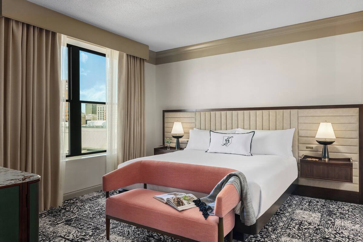 A hotel room with a large bed, two bedside tables, a chair, a window with curtains, and a patterned carpet.