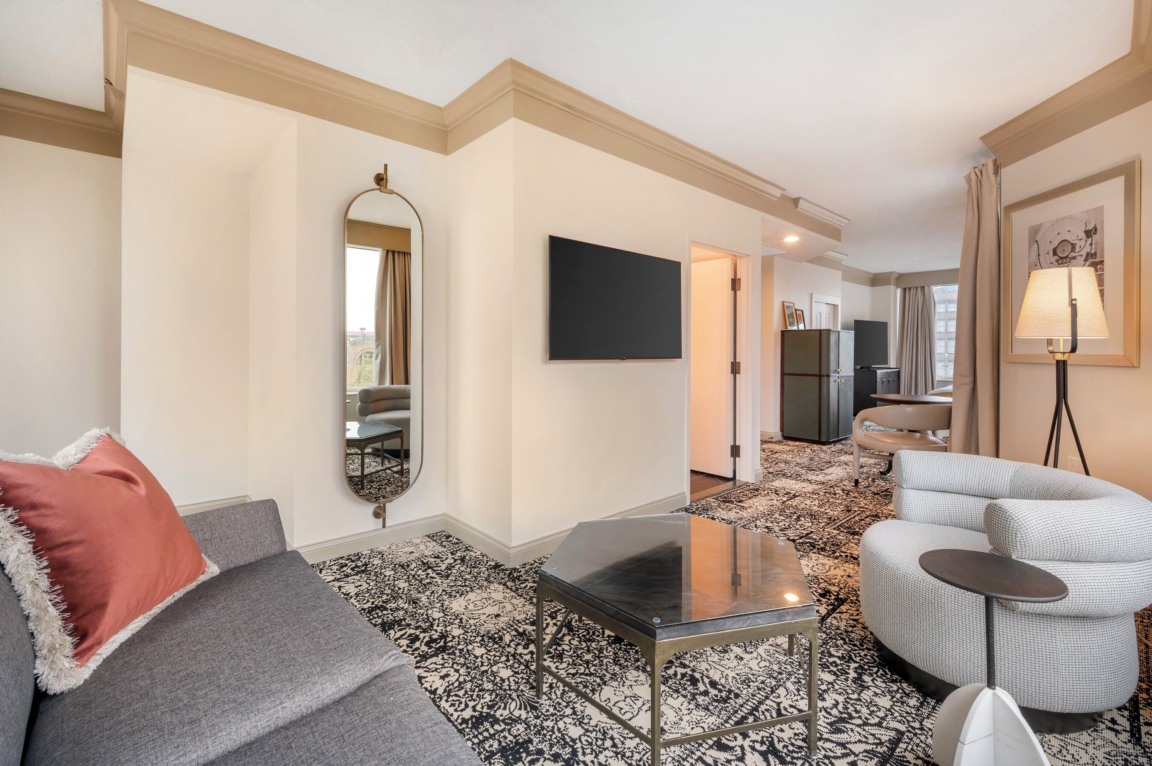 A modern hotel room with a grey sofa, a small black table, a white armchair, a large mirror, a flat-screen TV, and a patterned carpet.