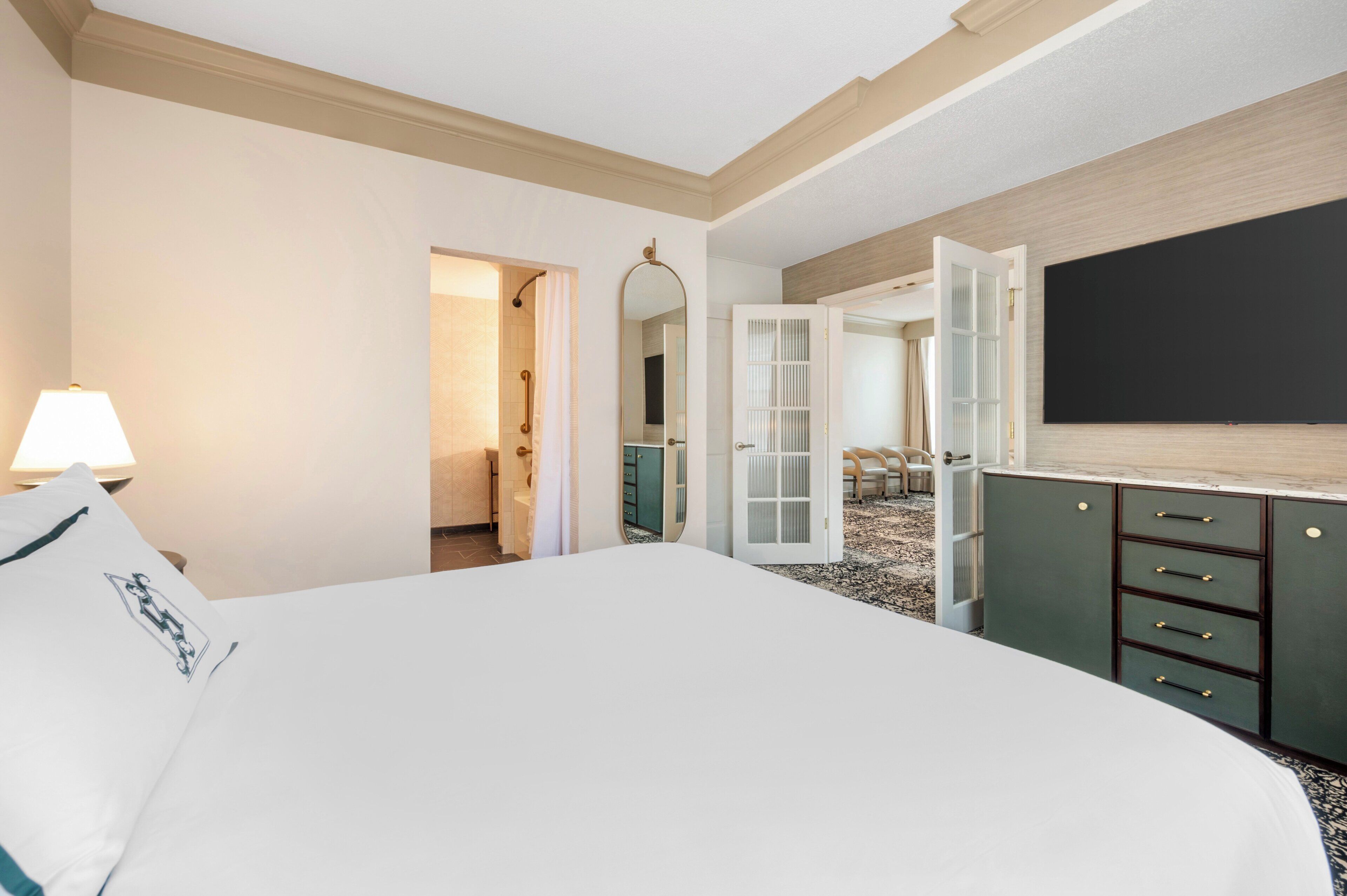 A hotel room with a large bed, a TV, a desk with drawers, a mirror, and a door leading to a bathroom.