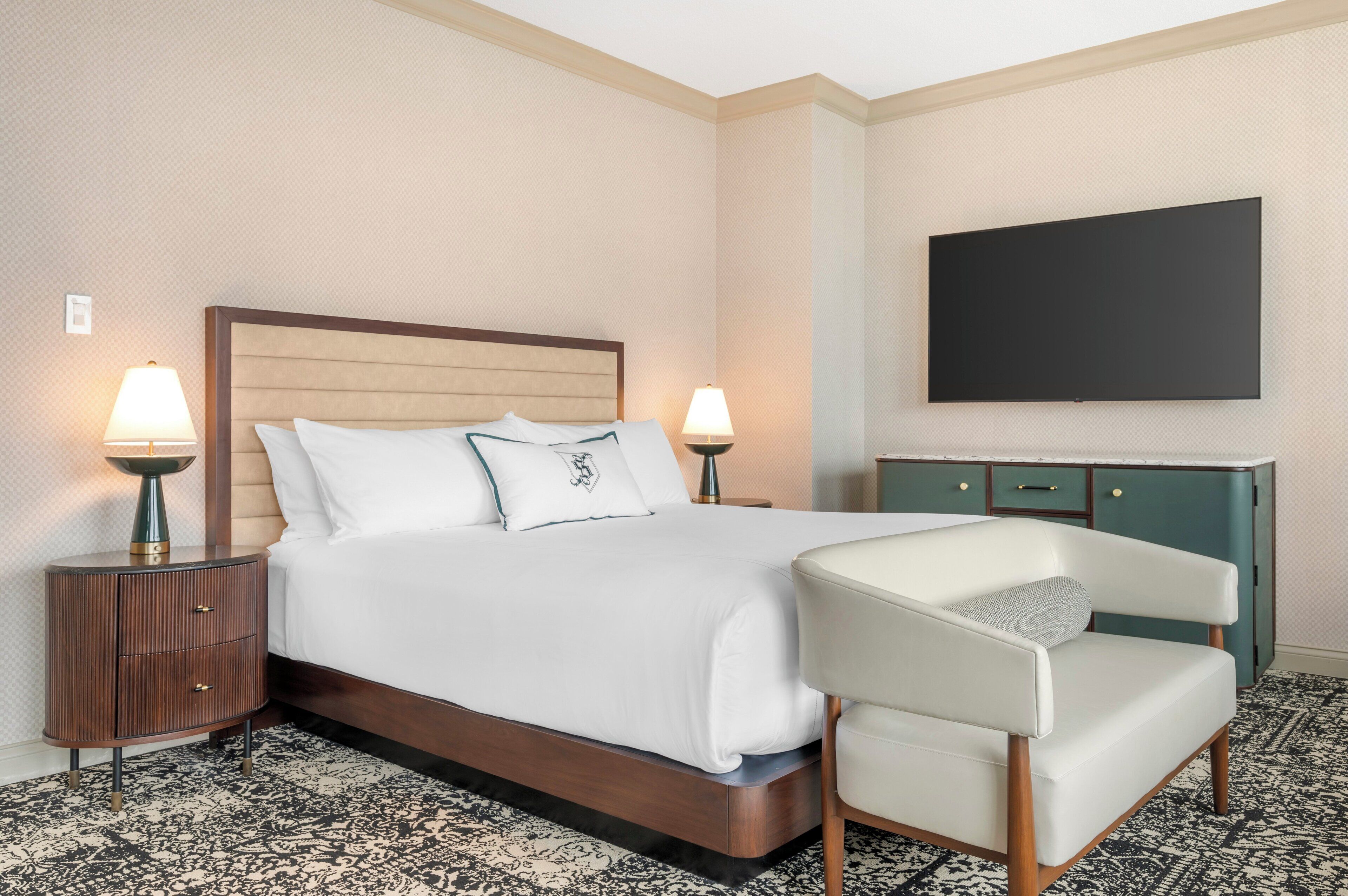 A hotel room with a large bed, two bedside tables with lamps, a flat-screen TV, a green dresser, and a white armchair.