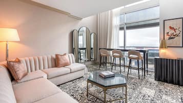 A modern hotel room with a sofa, chairs, a coffee table, and a large window offering a city view.