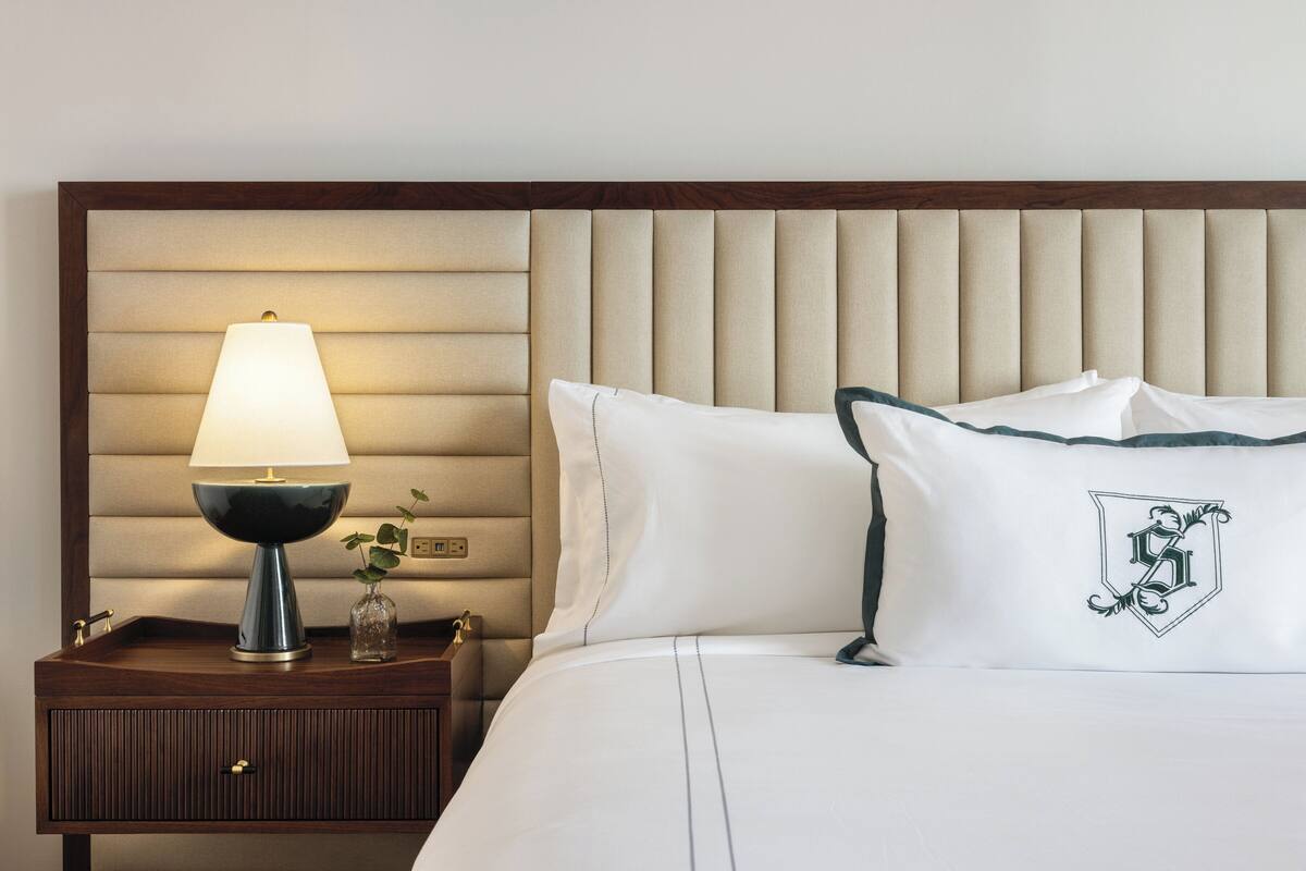 A neatly made bed with white linens and a pillow featuring a crest design. A bedside table with a lamp and a small vase of flowers is visible.