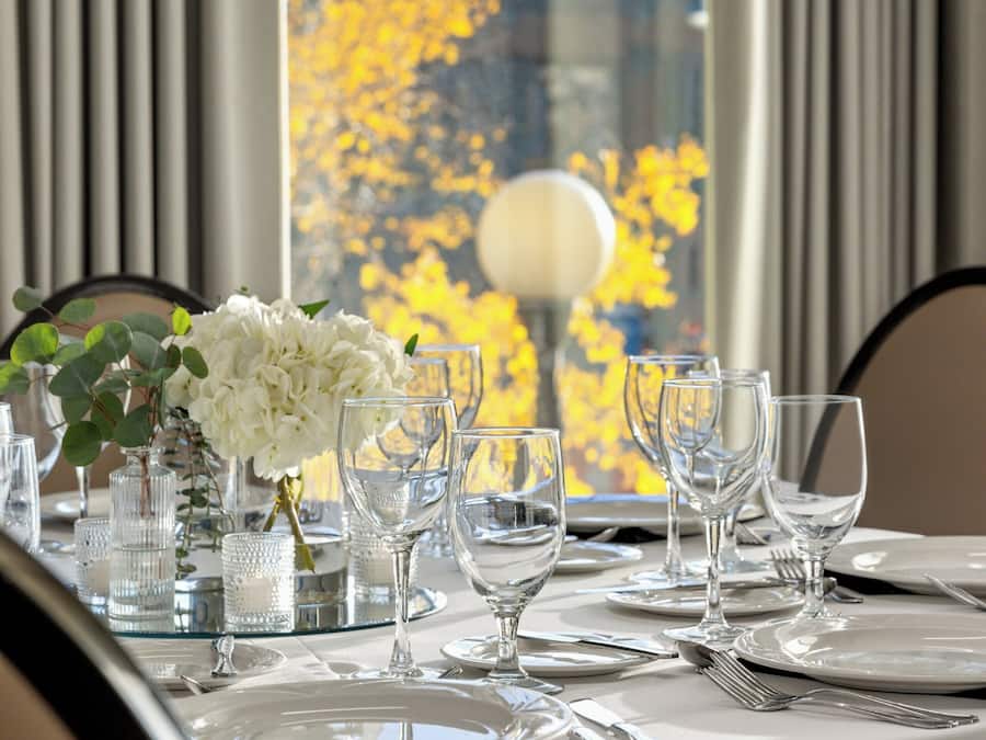 A dining table set with white plates, glasses, and cutlery, with a vase of white flowers and a candle holder.