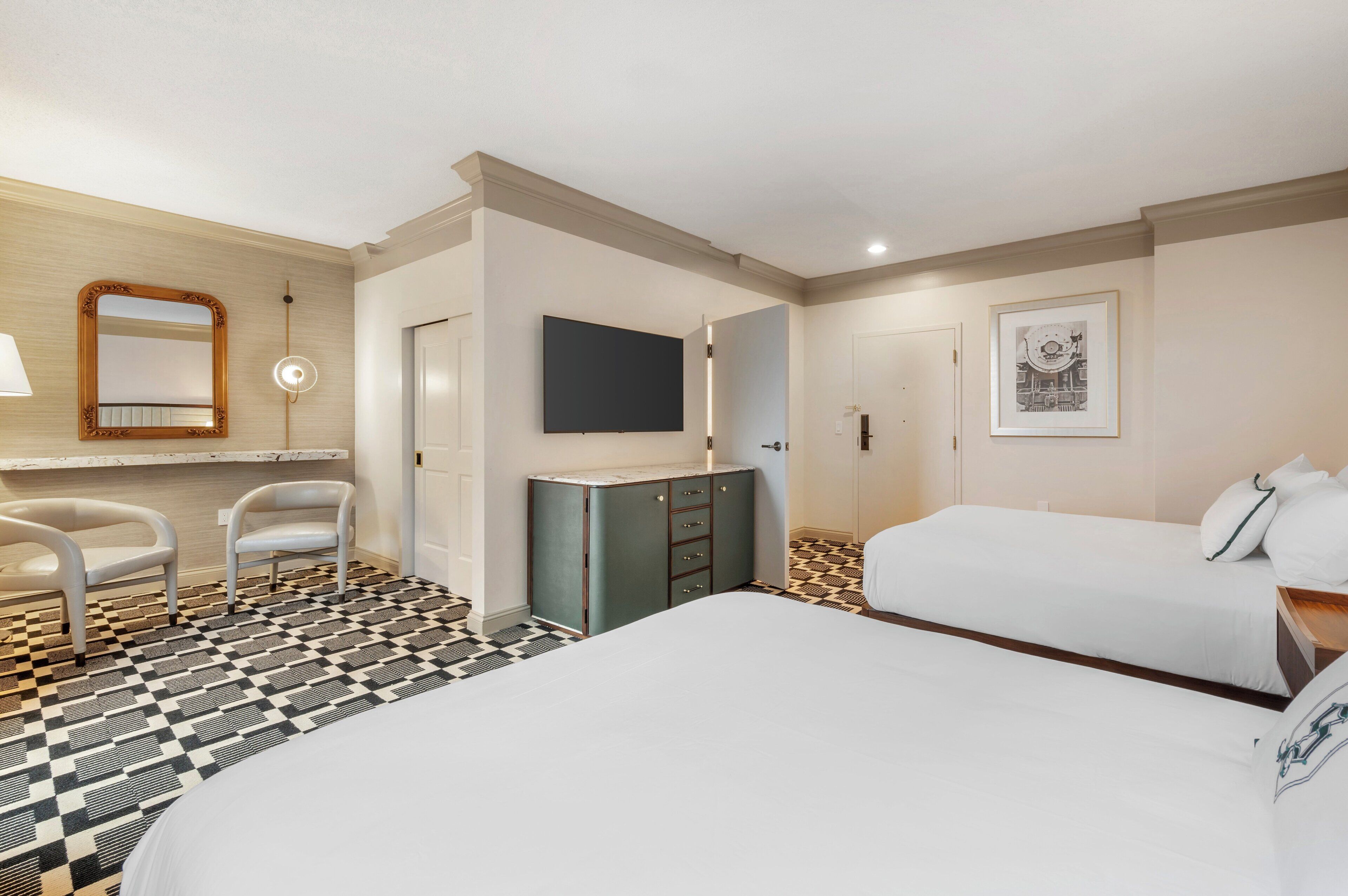 A hotel room with a checkered floor, a bed, a desk with a chair, a wardrobe, a mirror, and a flat-screen TV.